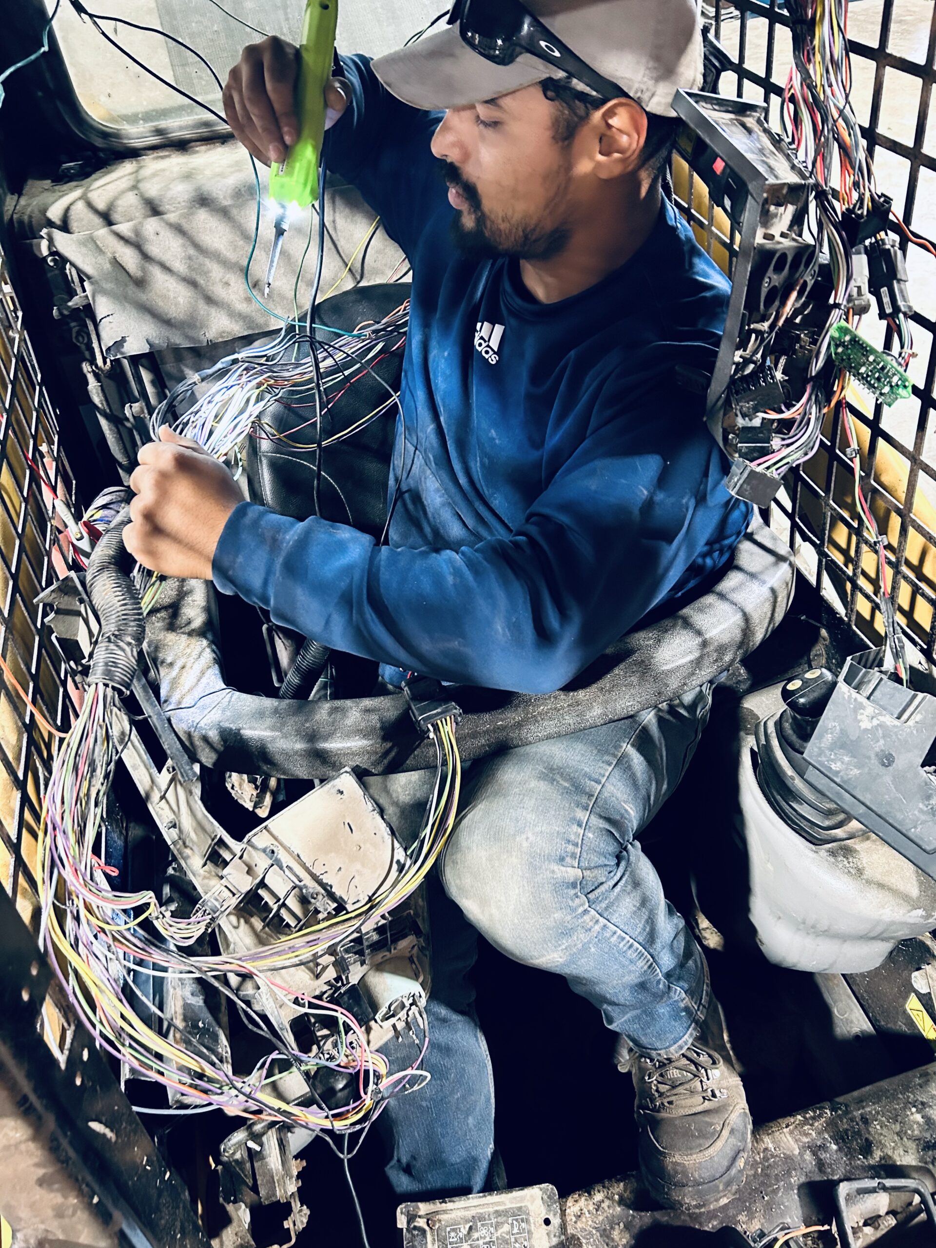 A technician works on electrical wiring inside a vehicle cab, using a flashlight to illuminate the complex array of colored wires and connectors.