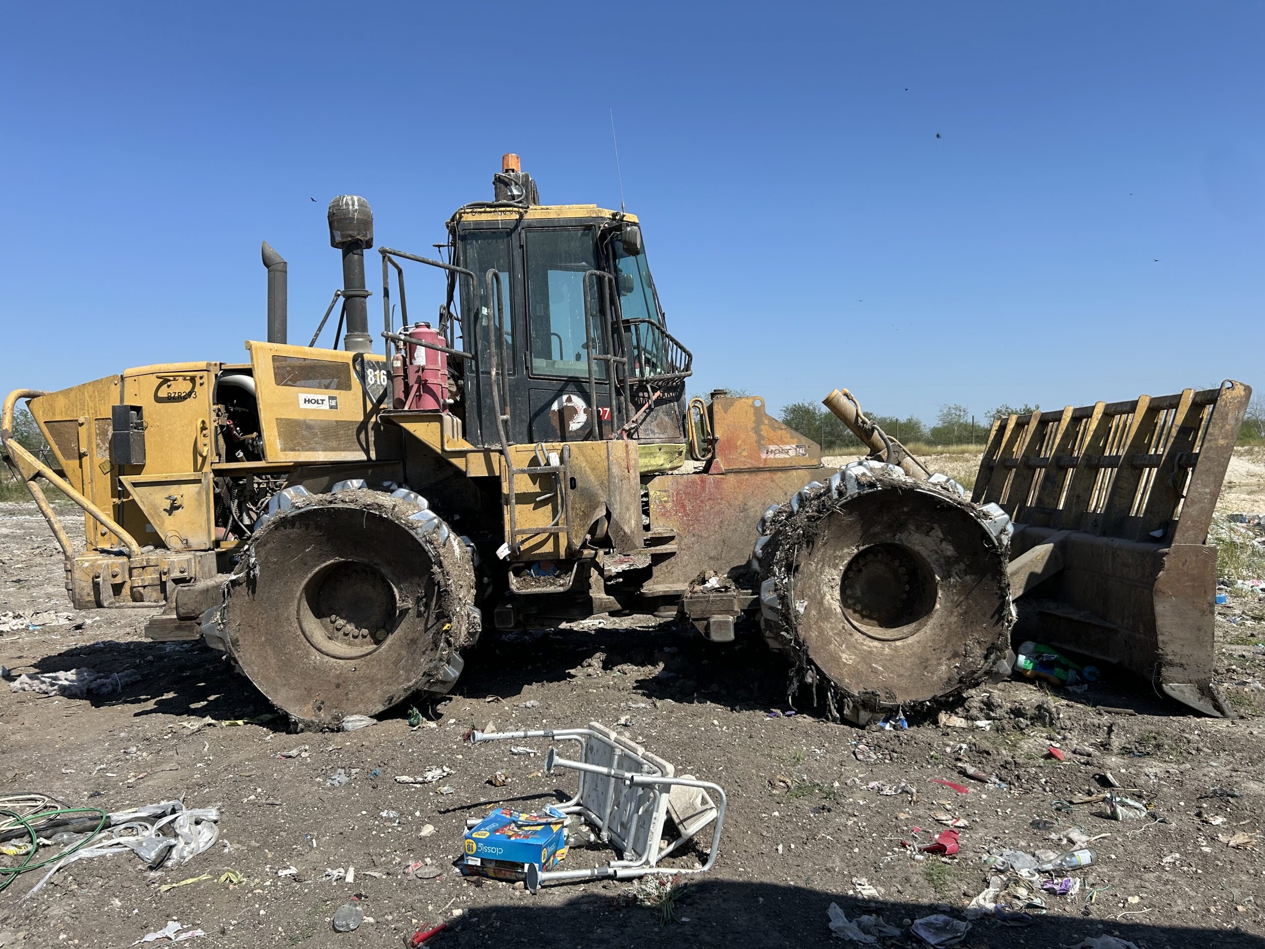 Heavy machinery, a yellow wheel loader, stands at a landfill site surrounded by debris. Its tires show signs of wear, highlighting the challenging working environment.