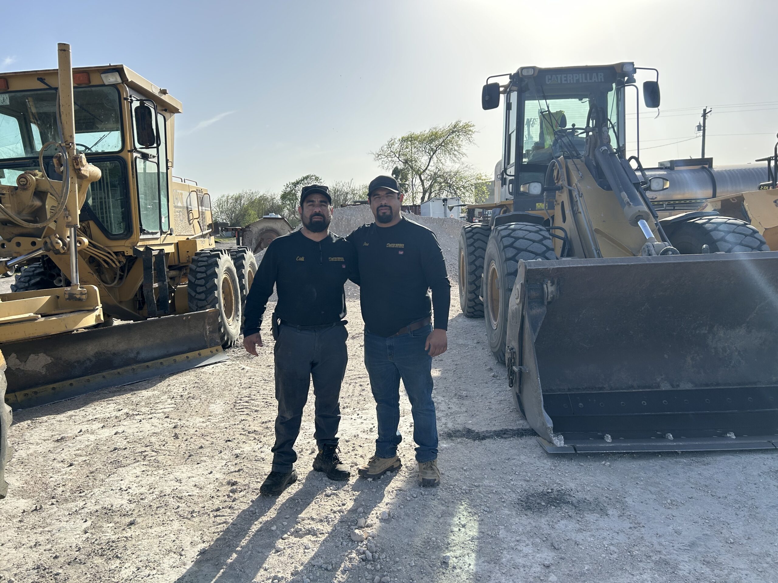 Two men stand together on a construction site, smiling, with heavy machinery, including a grader and loader, in the background.