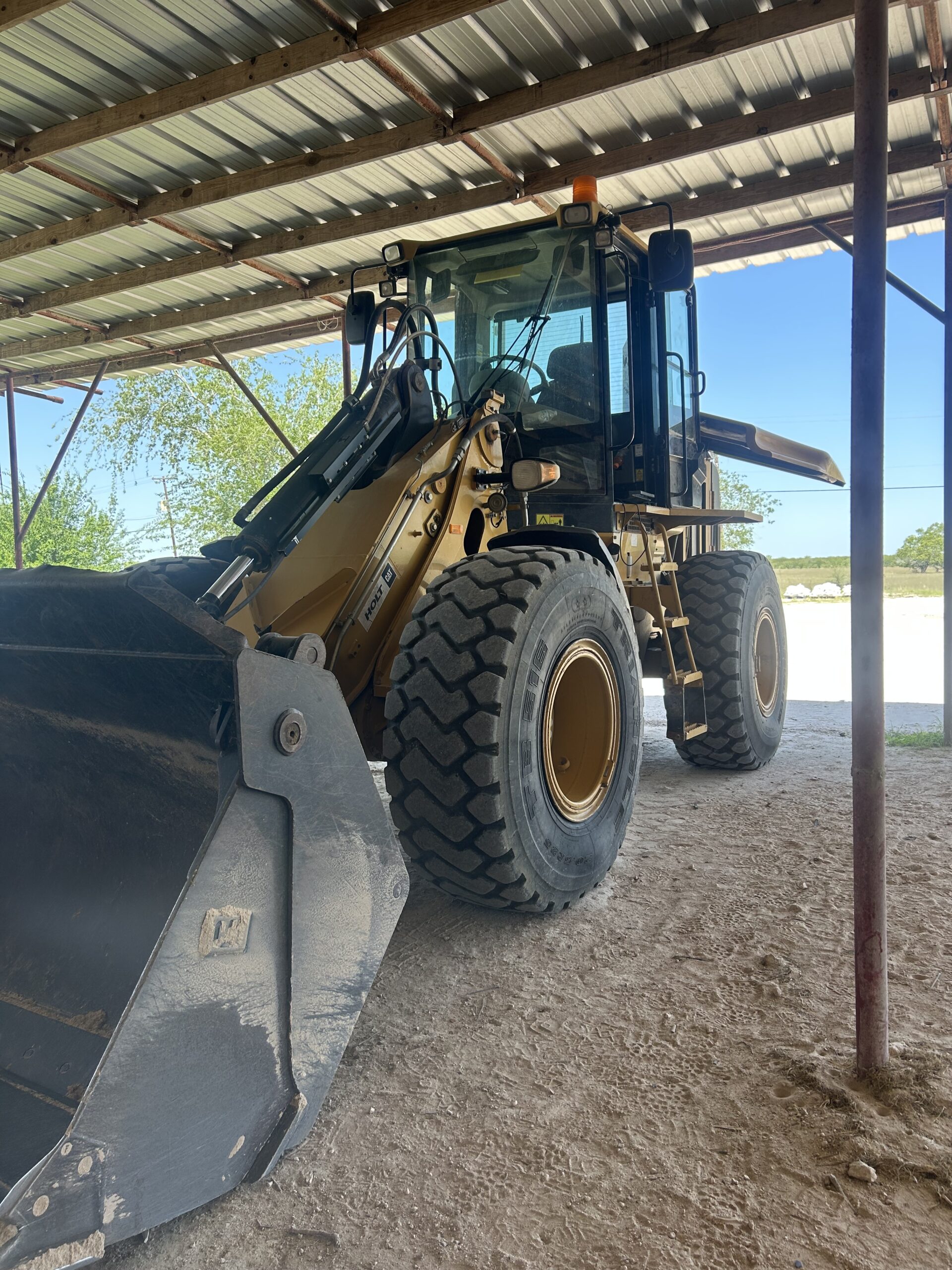 A front-end loader parked under a metal shed, showcasing its large tires and bucket; used for construction and landscaping tasks.