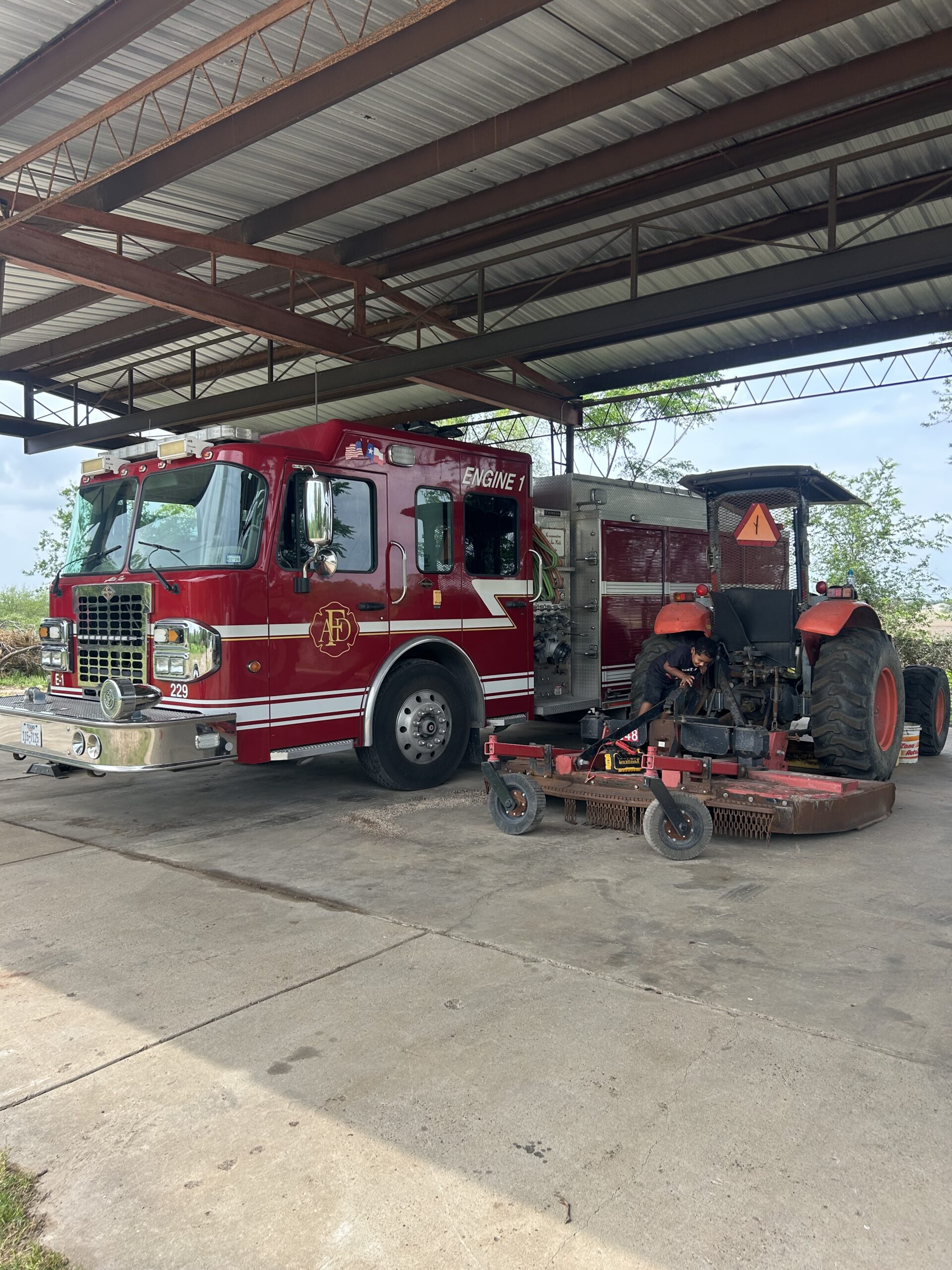 A fire truck labeled "Engine 1" is parked under a shelter, while a person works on a tractor's engine nearby, highlighting community services.