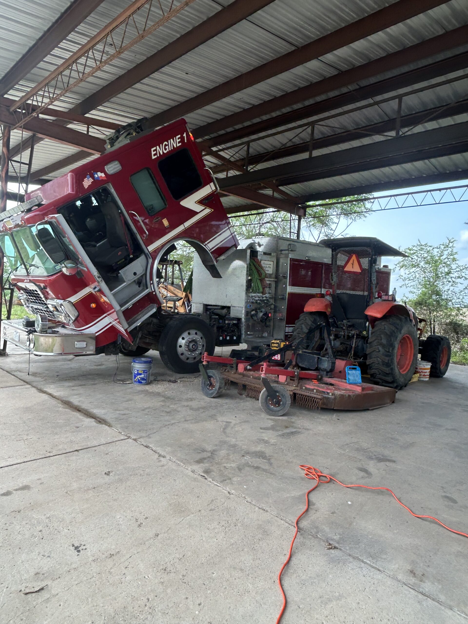 Fire truck engine raised for maintenance beside a red tractor and mowing equipment, in a sheltered workshop setting.