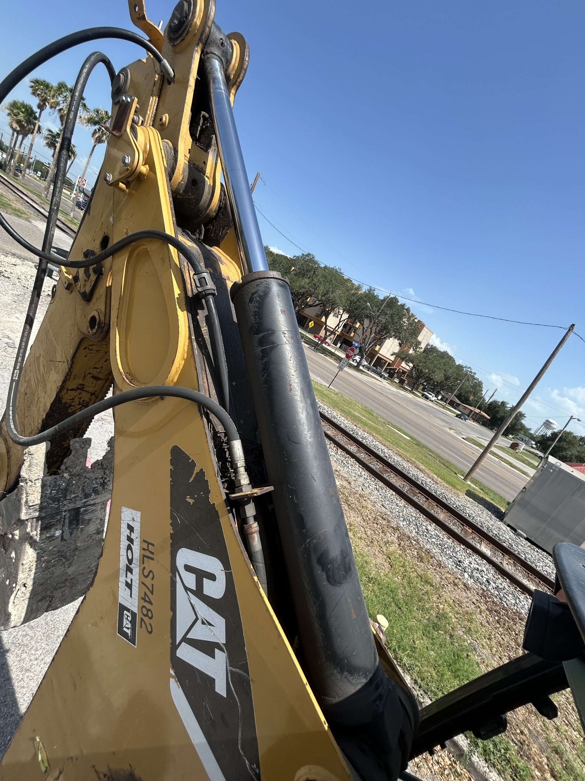 Close-up of a yellow Caterpillar backhoe loader's hydraulic arm, with a clear blue sky and road view in the background, highlighting construction equipment.